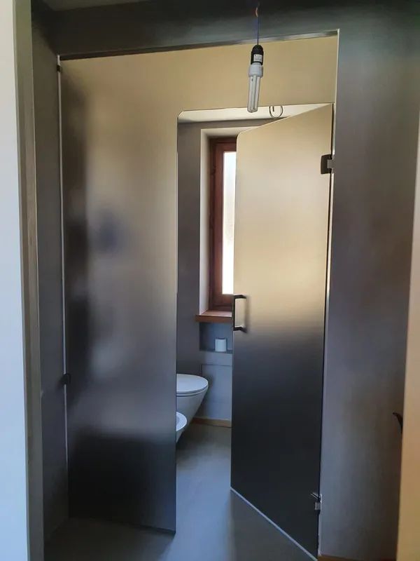 Cabina bagno aperta con porte grigio scuro e WC a vista all'interno, illuminato da una lampadina sospesa.
