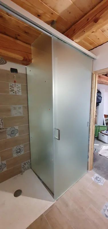 Box doccia in vetro in fase di costruzione in un bagno con pareti piastrellate e soffitto in legno.