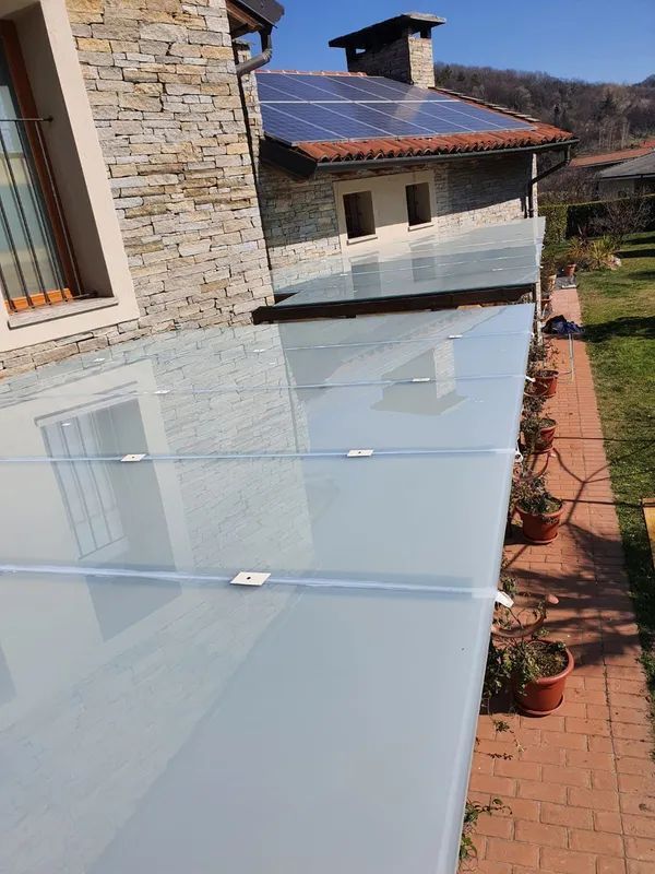 Patio coperto in vetro accanto a una casa in pietra, con vialetto in mattoni rossi e piante in vaso.