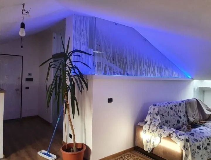 Accogliente soggiorno mansardato con parete bianca inclinata, illuminazione d'accento blu, divano, pianta in vaso e lampadina a sospensione.