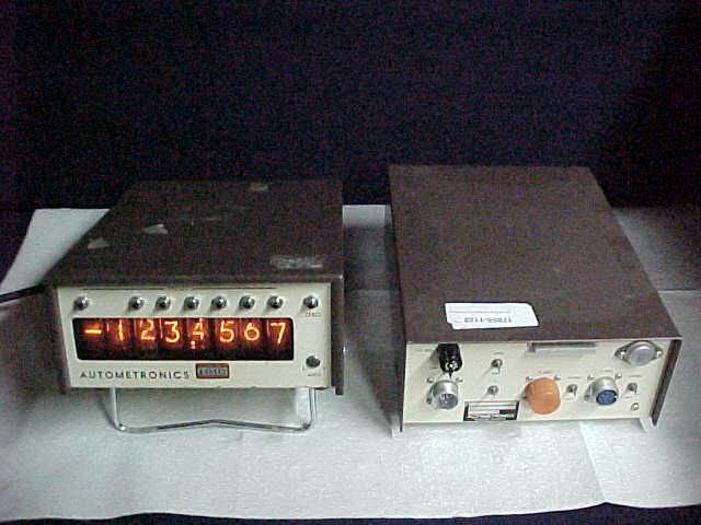 Two vintage electronic devices on a table, one with a lit numeric display and one with knobs and dials