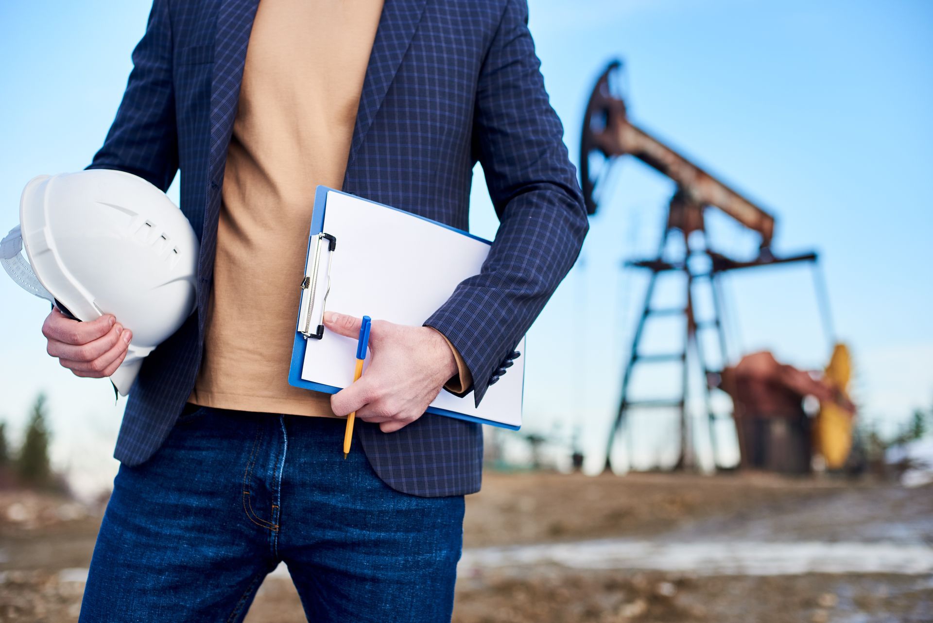 A man is holding a hard hat and a clipboard in front of an oil pump.