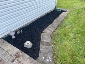 Mulched garden bed beside a white building, bordered by pavers and lawn, with small ornaments.
