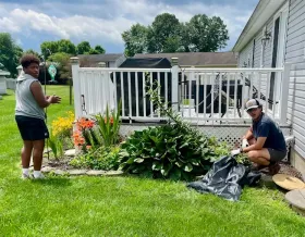 Two people gardening beside a house, planting flowers in a sunny backyard.