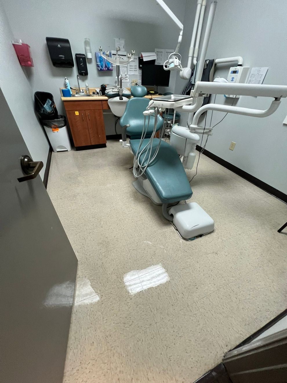 Dental exam room with teal chair, X-ray arm, sink, cabinets, and countertop equipment.