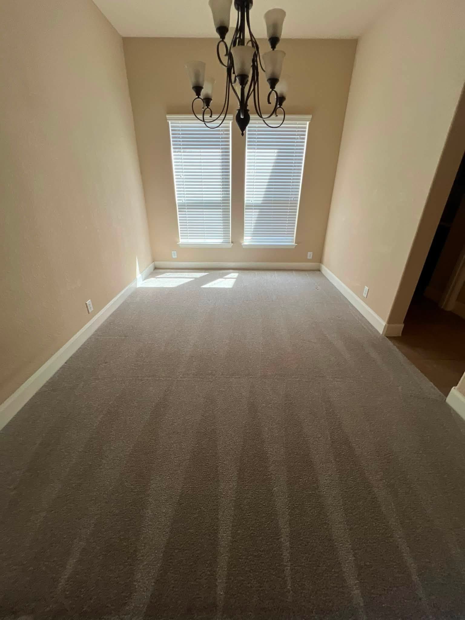Empty beige dining room with gray carpet, chandelier, and shuttered window