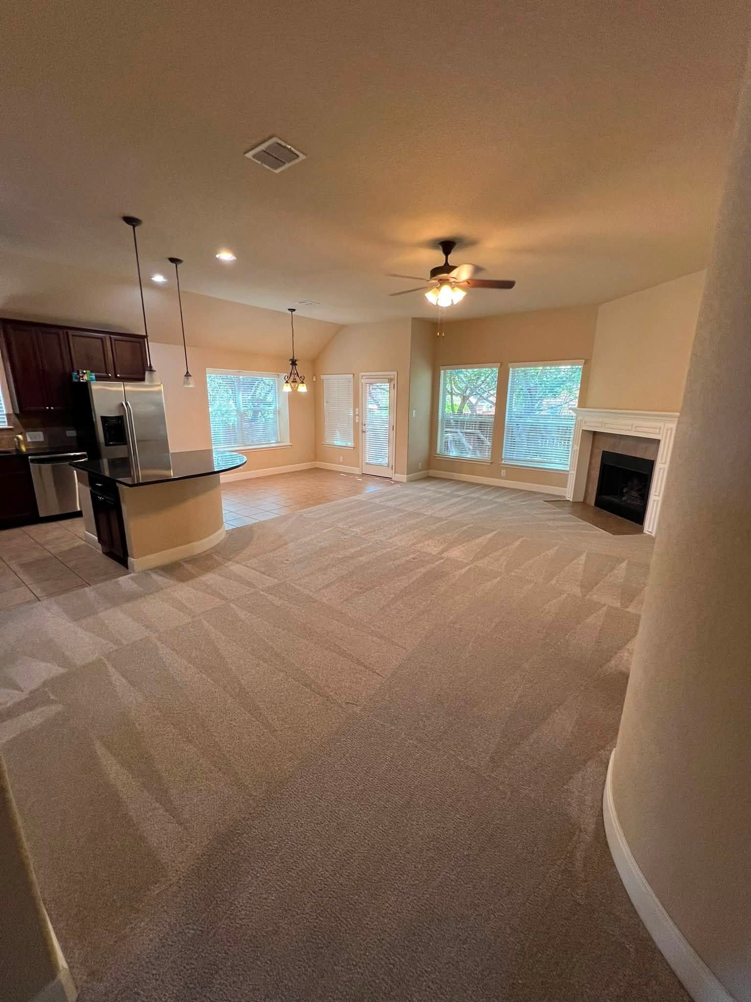 Empty carpeted living room with kitchen, fireplace, ceiling fan, and large windows.
