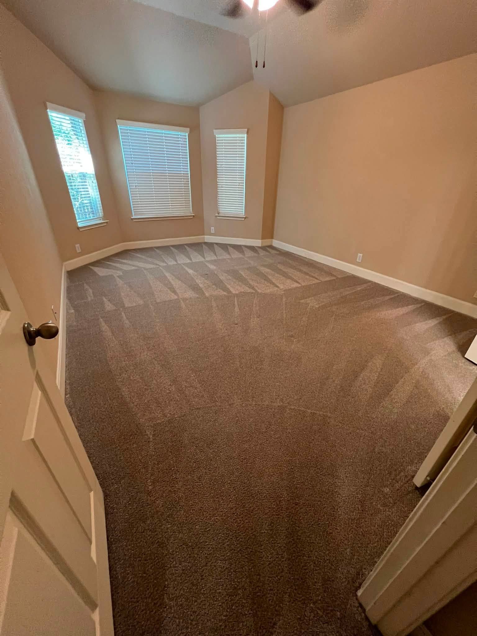 Empty carpeted bedroom with beige walls and windows showing trees outside.