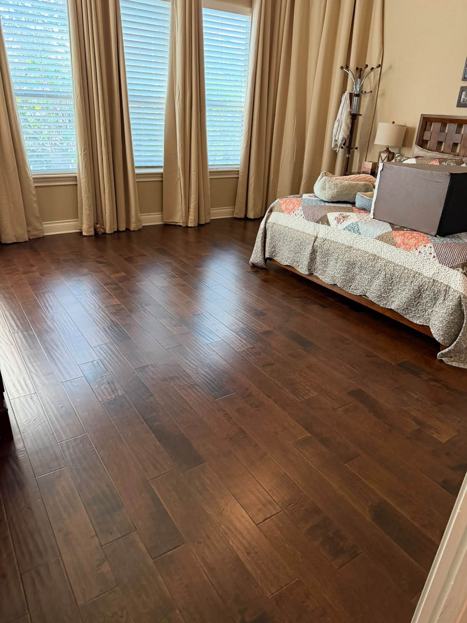 Empty bedroom with dark wood floors, beige curtains, and a bed with floral bedding and pillows.