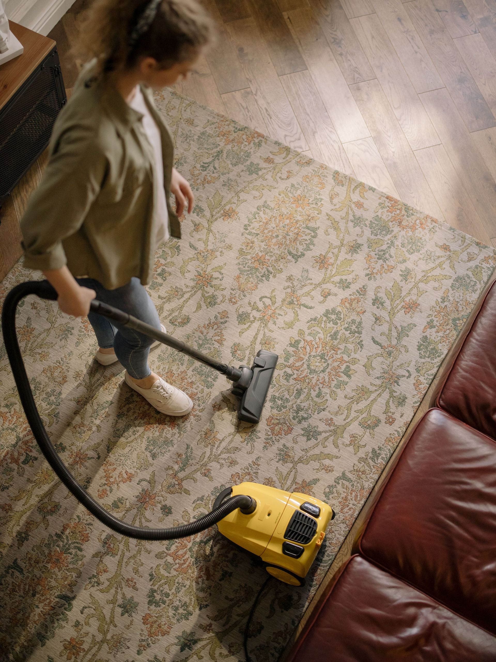 Person vacuuming a patterned rug beside a red couch with a yellow vacuum cleaner.