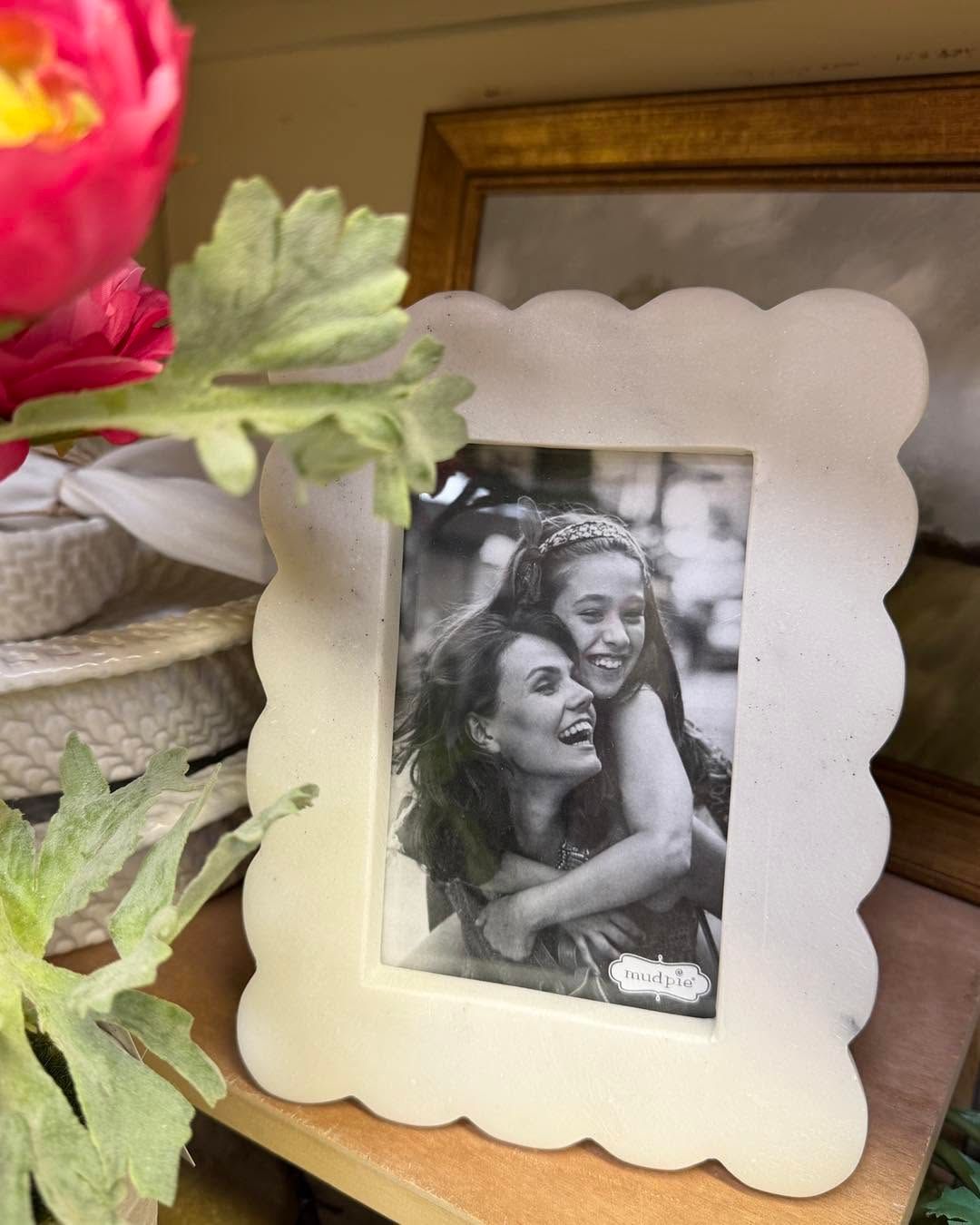 Black-and-white photo in a scalloped white frame on a shelf beside a pink flower and mirror