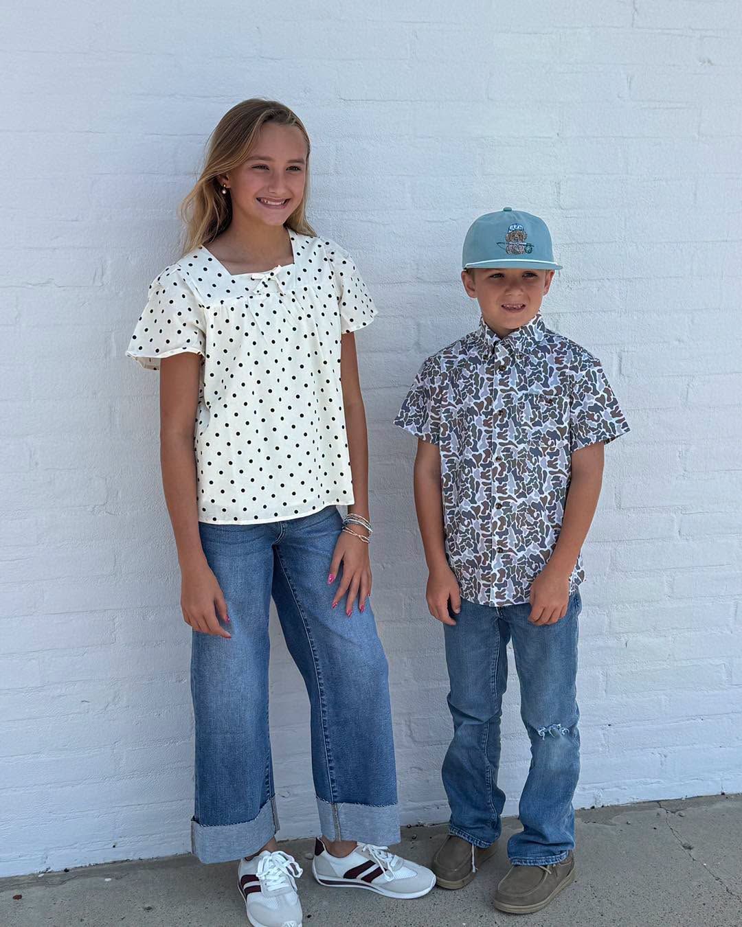Two children standing against a white wall, wearing patterned tops, jeans, and baseball caps.