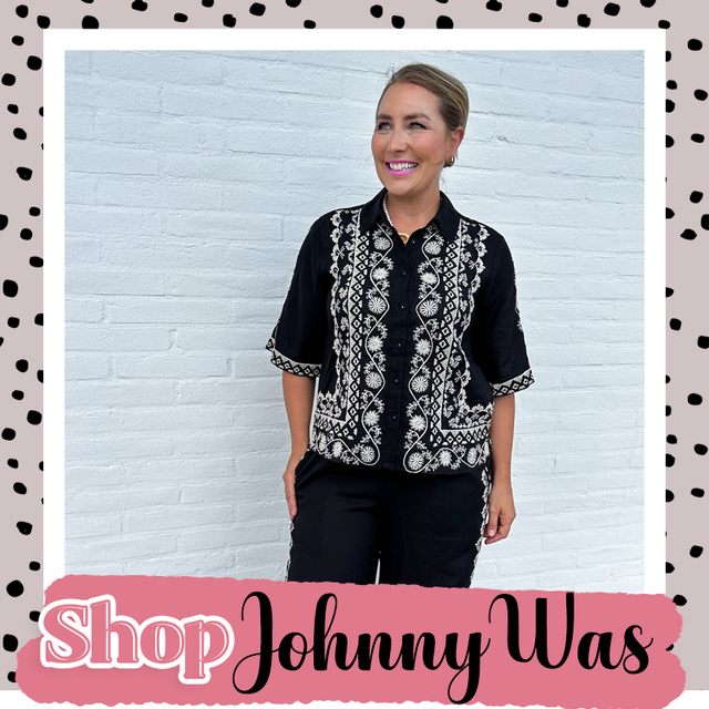 Woman modeling a black embroidered short-sleeve blouse against a white brick wall with Shop Johnny Was branding
