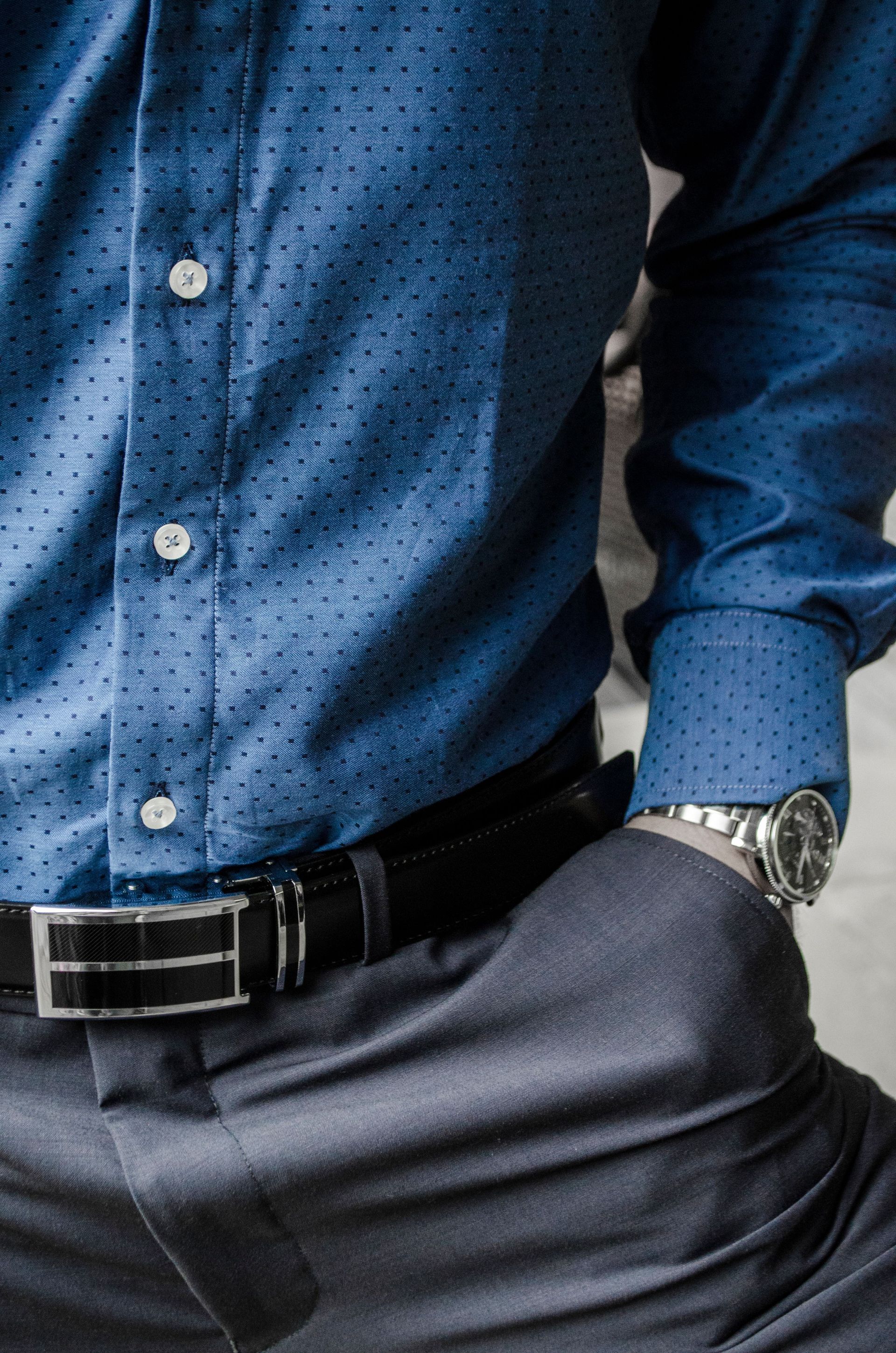 Man in blue patterned dress shirt with watch and black belt, hands in pockets, wearing dark trousers.
