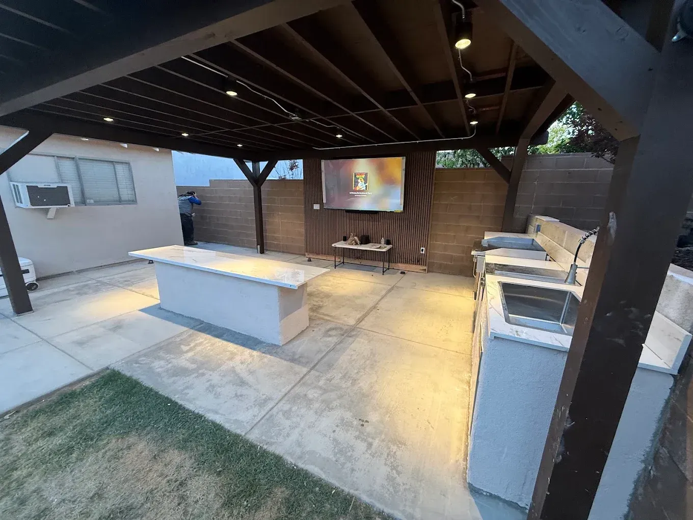 Covered backyard patio with grill, picnic table, projector screen, and concrete seating area at dusk