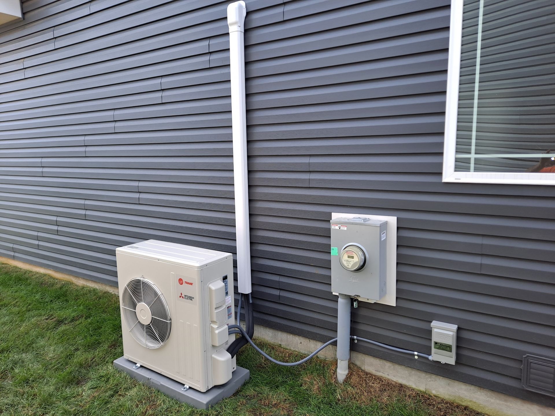 A white mini-split air conditioner is sitting on the side of a house next to a window.