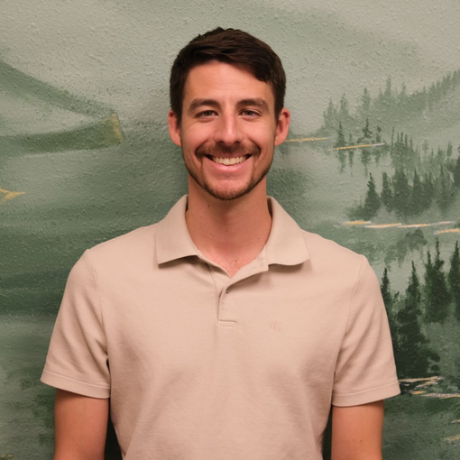 Smiling man in a beige polo shirt standing before a painted forest and lake backdrop