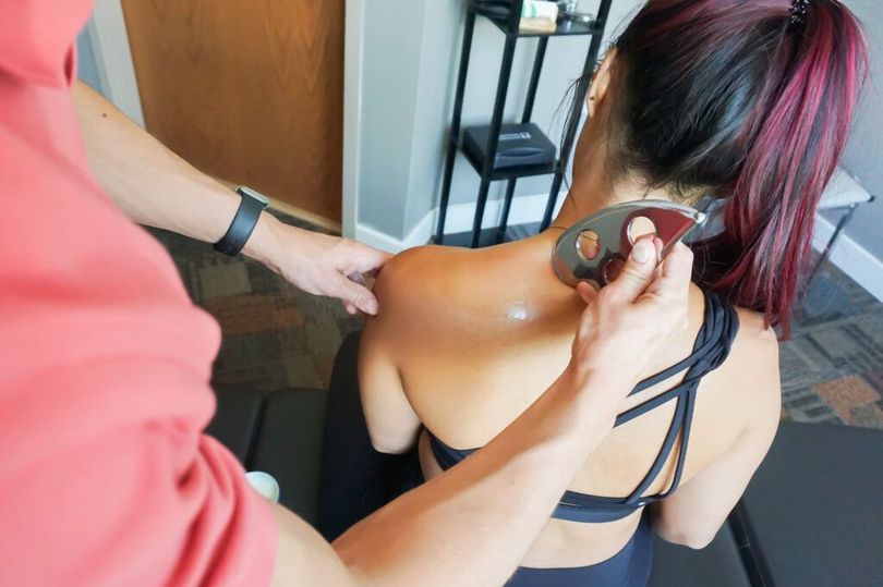 Person undergoing a shoulder massage in a salon, with another person using a massage tool on the upper back.