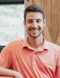 Smiling man in a coral polo shirt leaning by a window with wooden paneling behind him