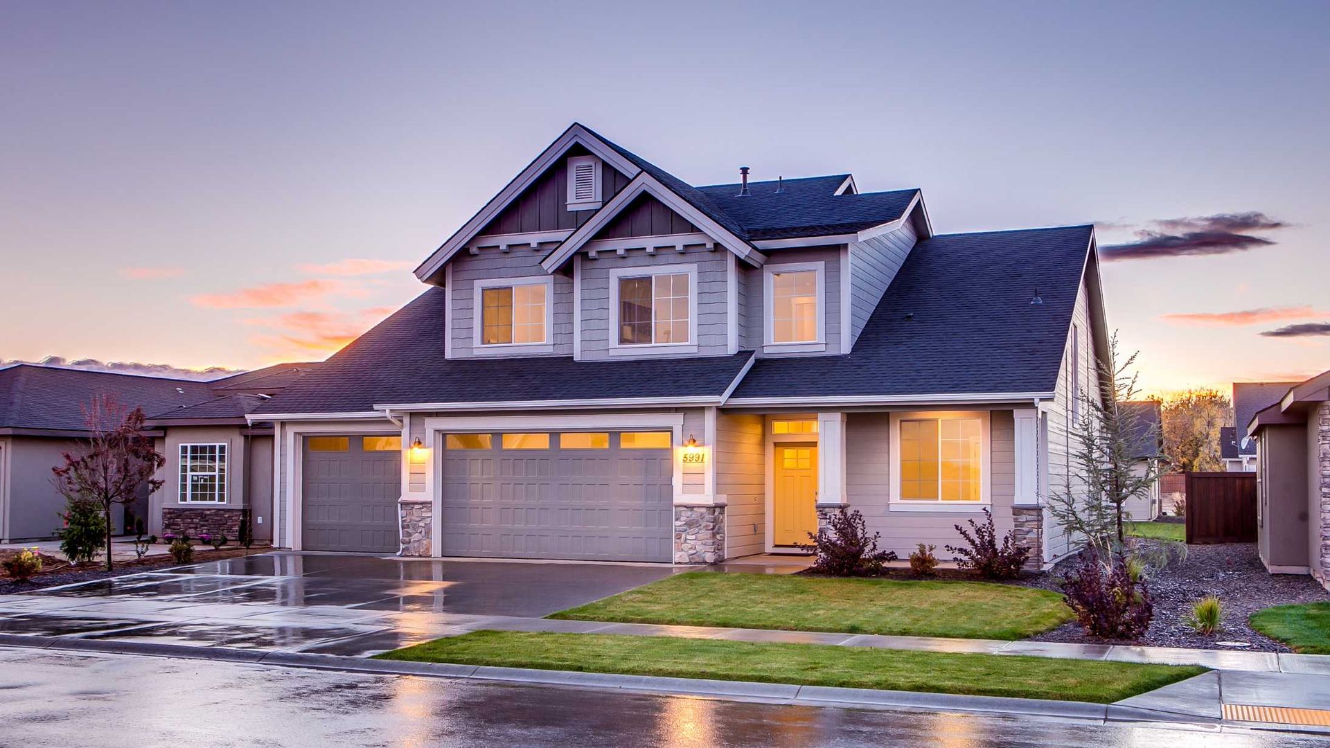 Suburban house at dusk with lit windows, wet driveway, and a pink sunset sky