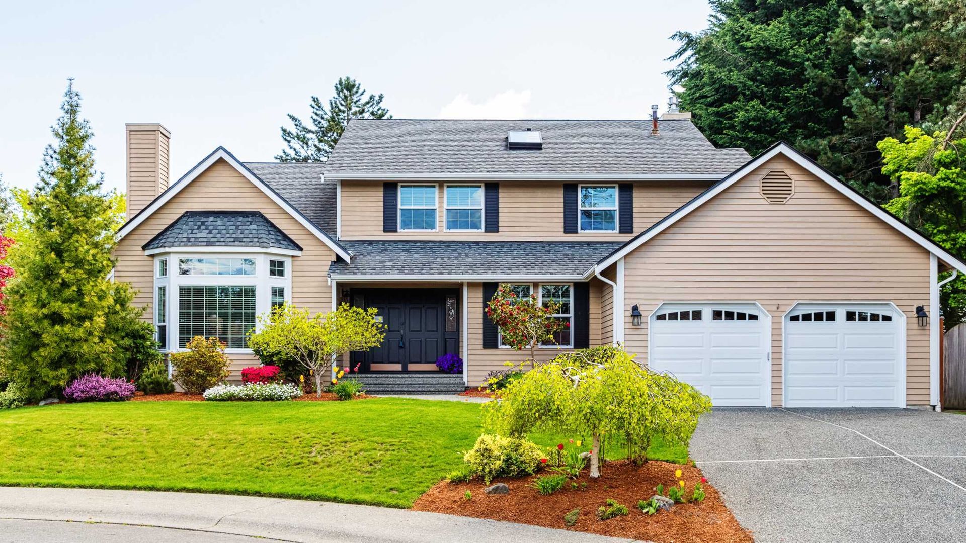 Suburban house with stone and beige siding, front lawn, flower beds, and a two-car garage
