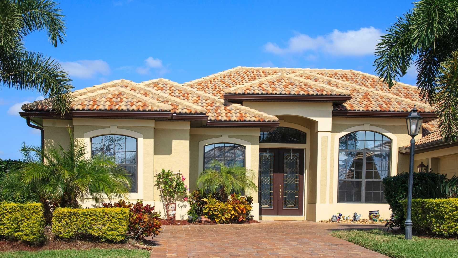 Single-story cream house with a tiled roof, arched windows, and a landscaped front yard under a blue sky