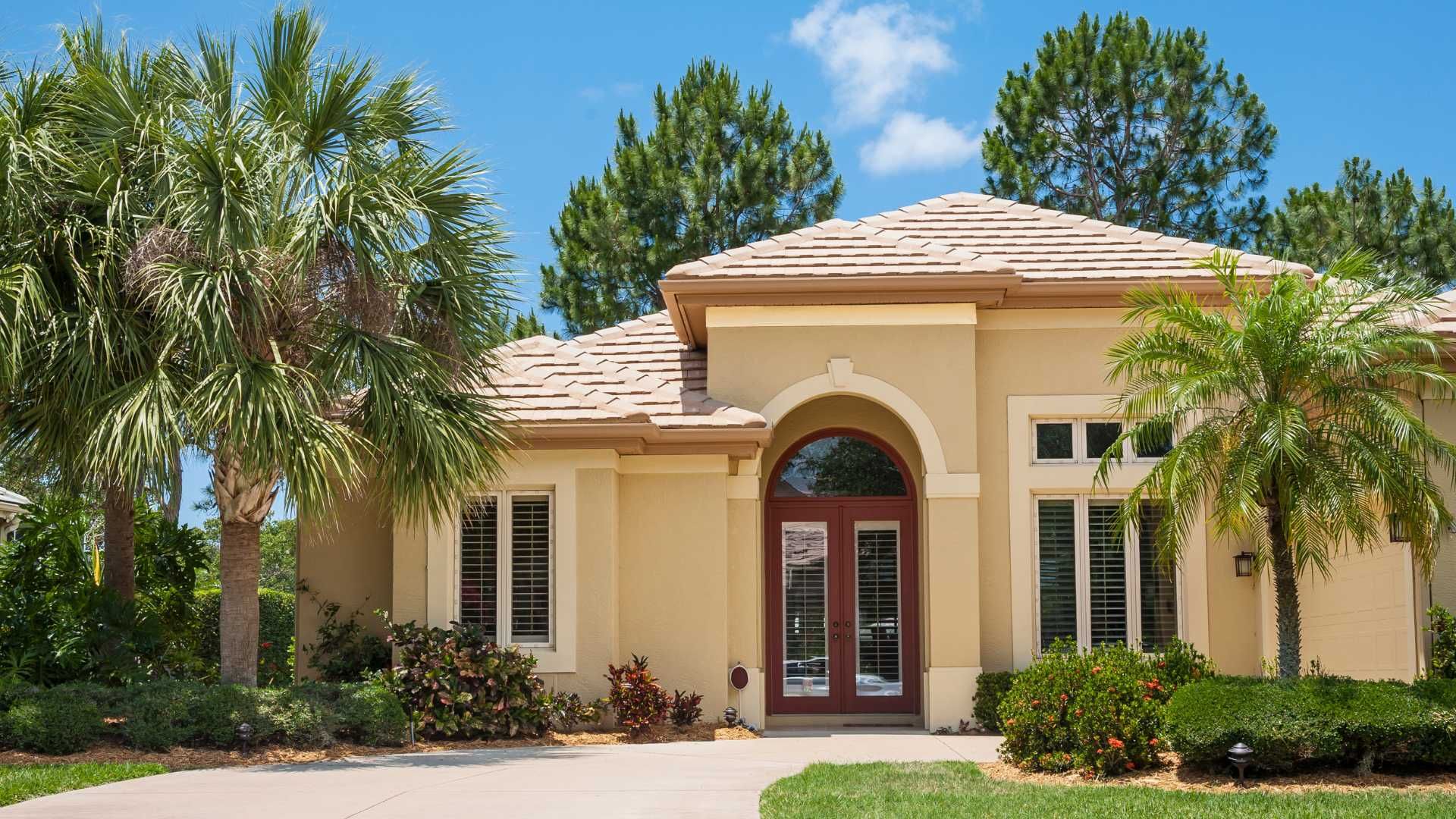 Single-story beige house with a red arched front door, palm trees, and a sunny landscaped yard