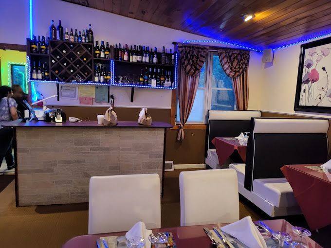 Restaurant interior with bar, wine rack, booths, tables with red tablecloths, and blue lighting.