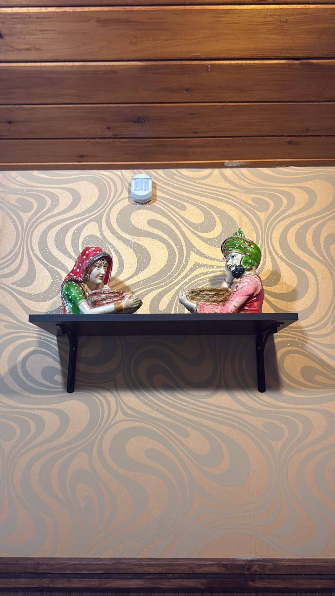 Shelf with two ceramic figures, woman in red scarf and man in green hat seated, ornate wallpaper.
