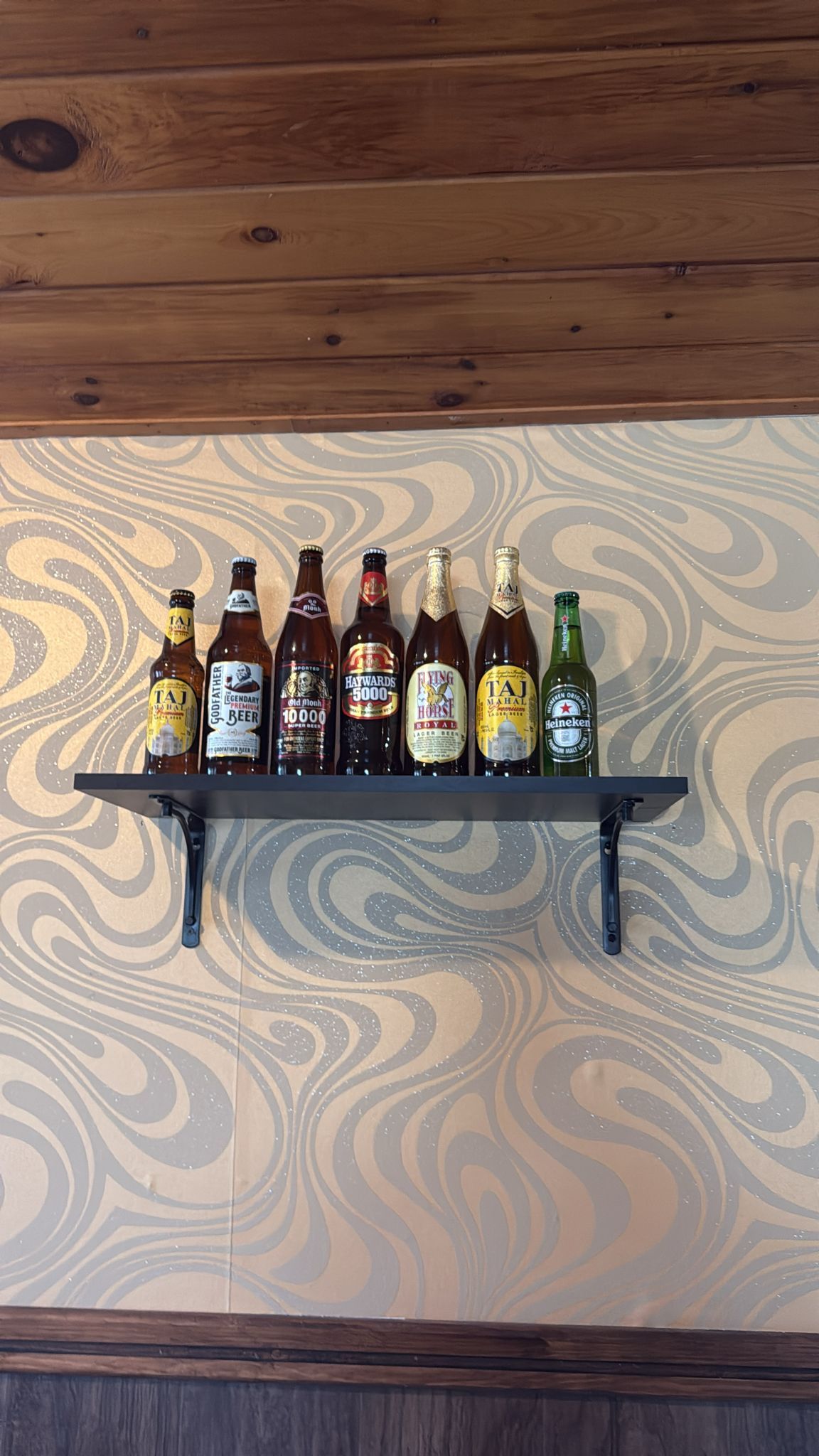 Shelf with eight beer bottles against a gold and silver patterned wallpaper and wood trim.