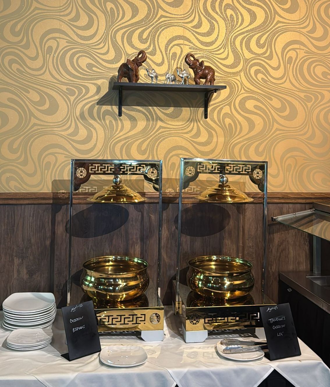 Two golden buffet chafing dishes on a table with a stack of plates. Beige and gold swirled wallpaper.