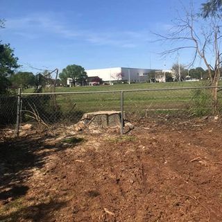 A chain link fence surrounds a dirt field with a large building in the background.