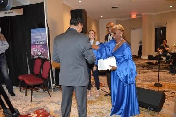 Two people in formal attire exchanging an award in a hotel ballroom setup.