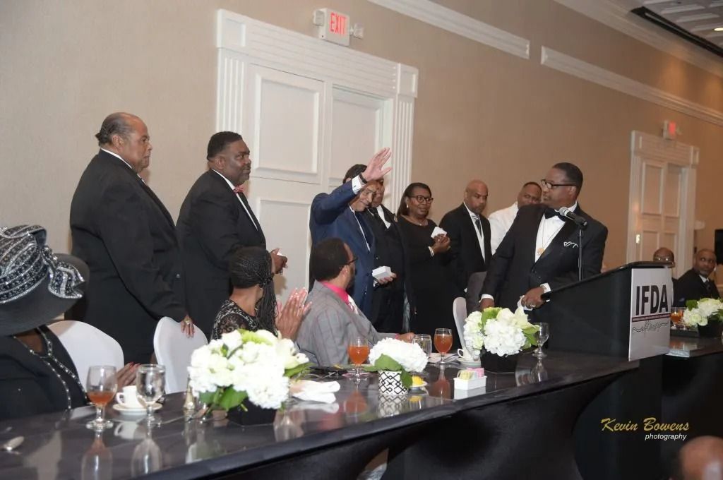 Formal banquet scene with people standing and seated around a long table, one man raising his hand in a toast