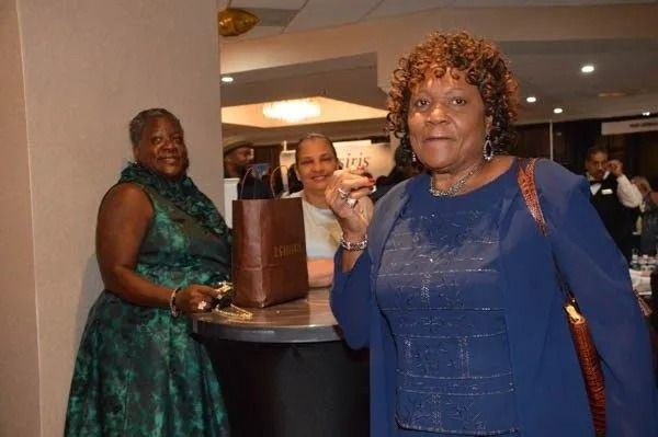 Three women at a social event, one in a blue dress holding a drink in the foreground.