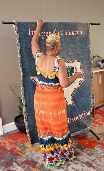 Woman in orange dress raising arms before a blue “Independent Funeral Association” backdrop