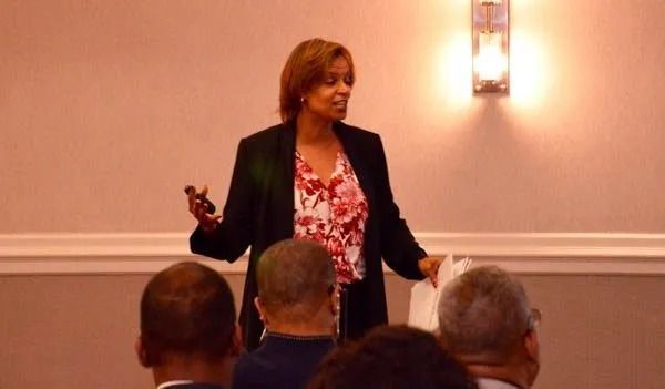 Speaker presenting in a conference room, standing by a wall light with audience seated in foreground.