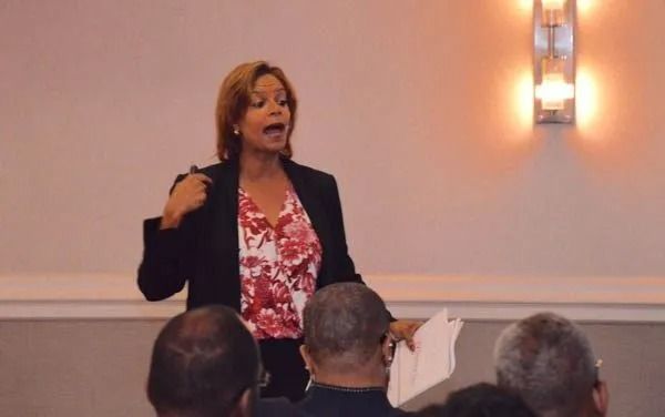 Woman speaking at a podium in a conference room, gesturing with papers in hand