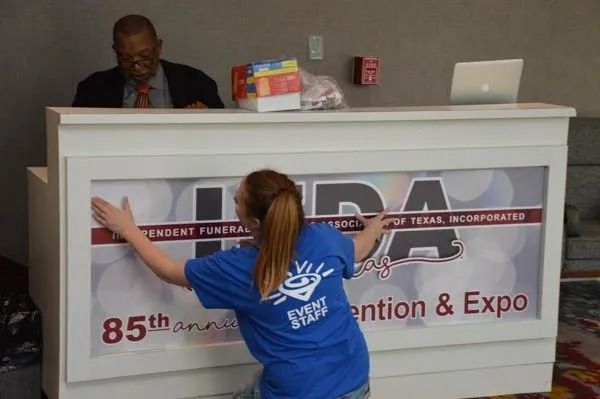 Woman in blue shirt leans on expo booth counter while man stands behind it with a laptop.