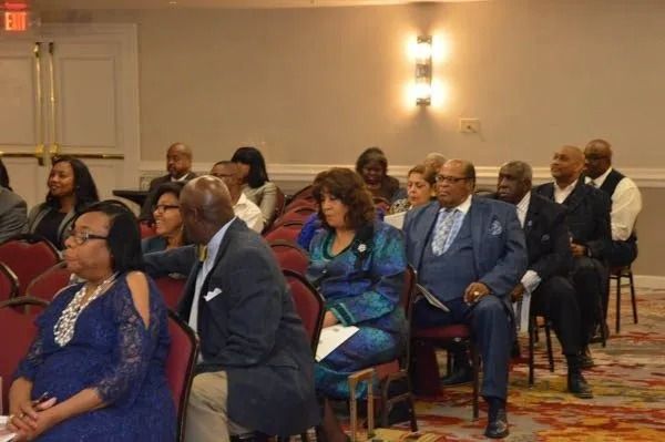 Audience seated at a formal indoor event, facing forward in red chairs.