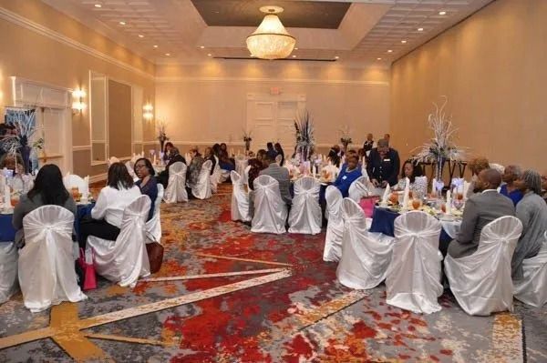 Banquet hall with guests seated at round tables, white chair covers, and warm ceiling lights on a patterned carpet.