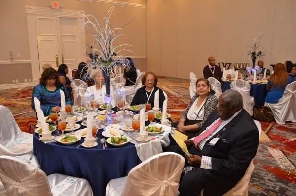 Formal banquet guests seated at round tables in a banquet hall with blue tablecloths and floral centerpieces