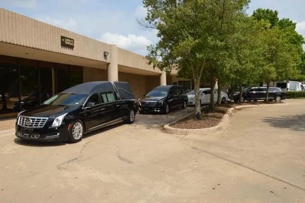 Black hearse and cars parked outside a building with trees and a dirt driveway