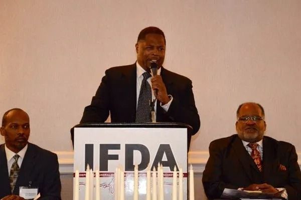 Man speaking at a podium with IFDA sign, flanked by two seated men in suits
