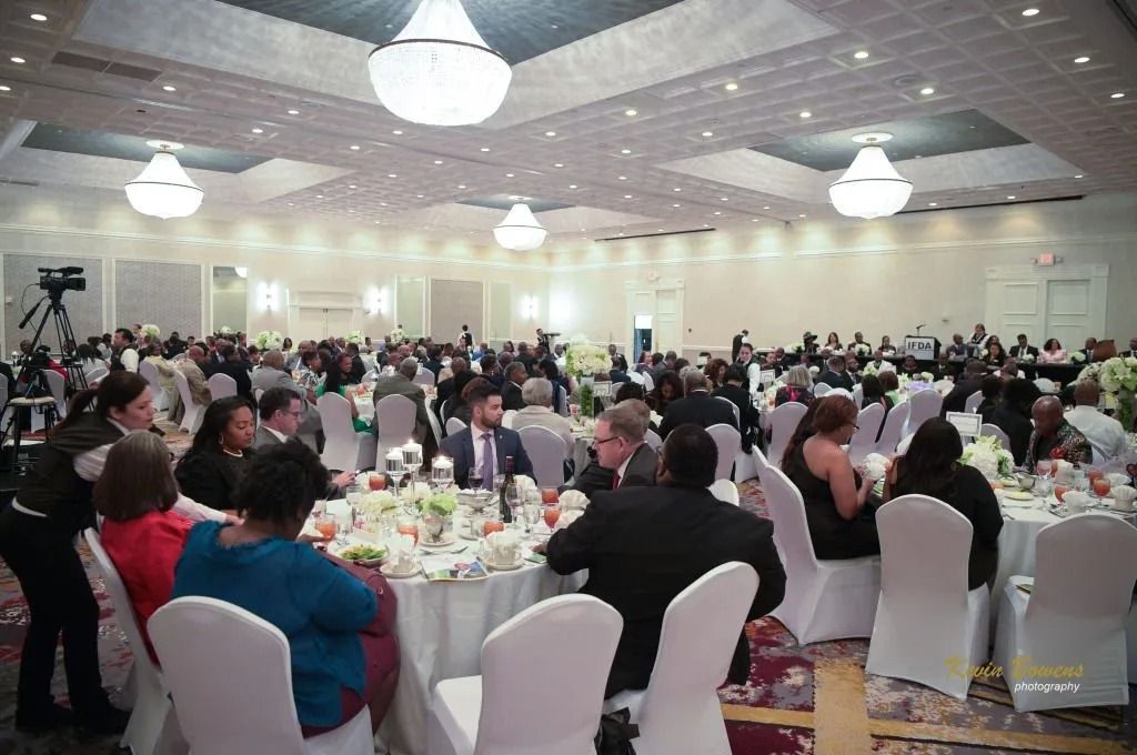 Crowded banquet hall with round tables, seated guests, and white chair covers under hanging lights