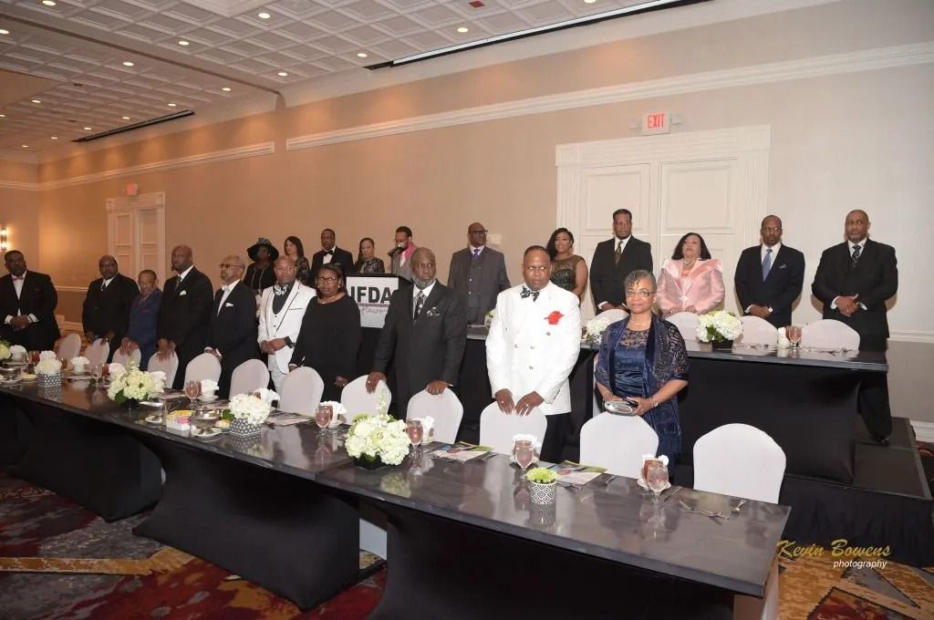 People standing at a formal banquet in a hotel ballroom, with tables set in front of them.