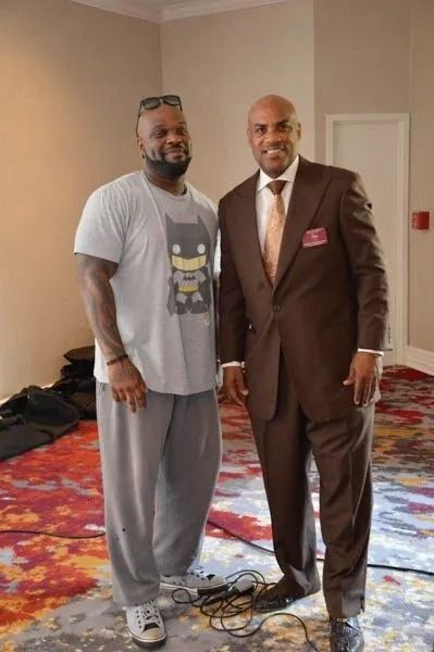 Two men standing together indoors on a patterned carpet, one in gray casual clothes and one in a brown suit.