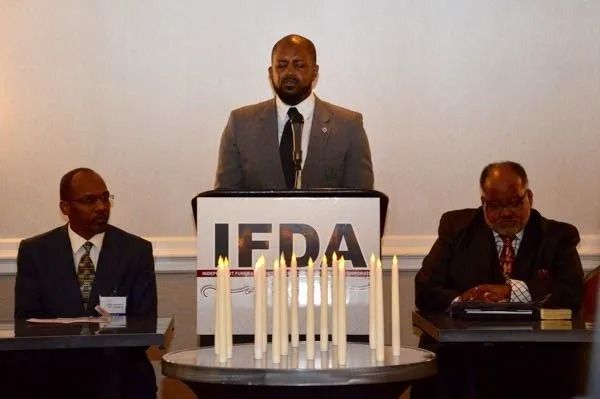 Three men at a podium with “JFDA” sign, two seated beside him in a formal meeting room.