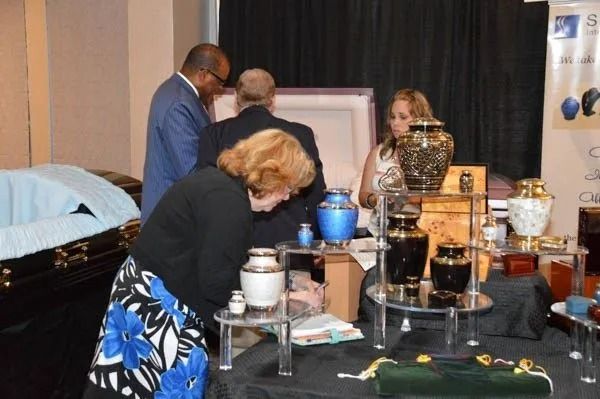 People gathered around a display table with bottles, cups, and a blue vase at an indoor event.