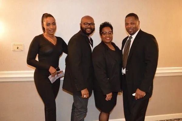 Four people in formal black attire posing together indoors against a beige wall.